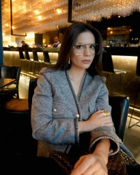 Woman in a grey tweed jacket wearing oversized gold glasses seated in a dimly lit restaurant bar