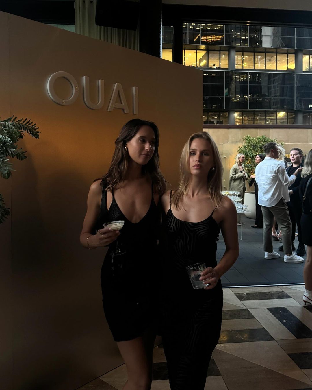 Two adult women in black dresses stand together indoors, holding drinks in glasses, with a neutral-toned wall and city buildings visible outside.