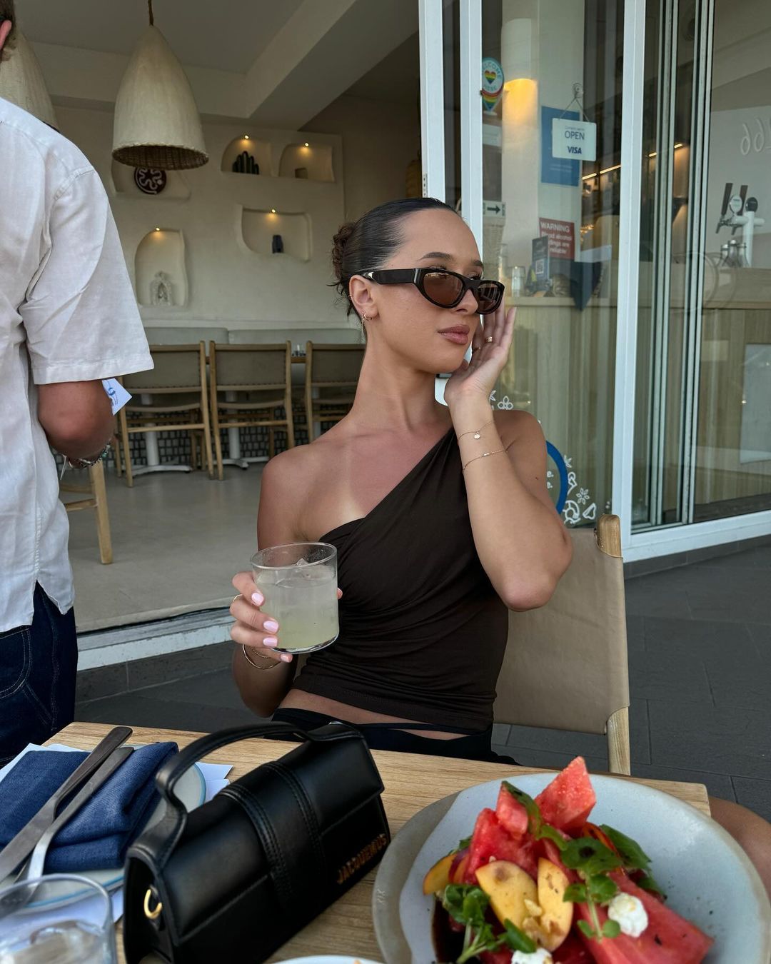 A brunette woman in sunglasses and a brown top sits at an outdoor table with a drink, a salad, and a black Jacquemus purse.