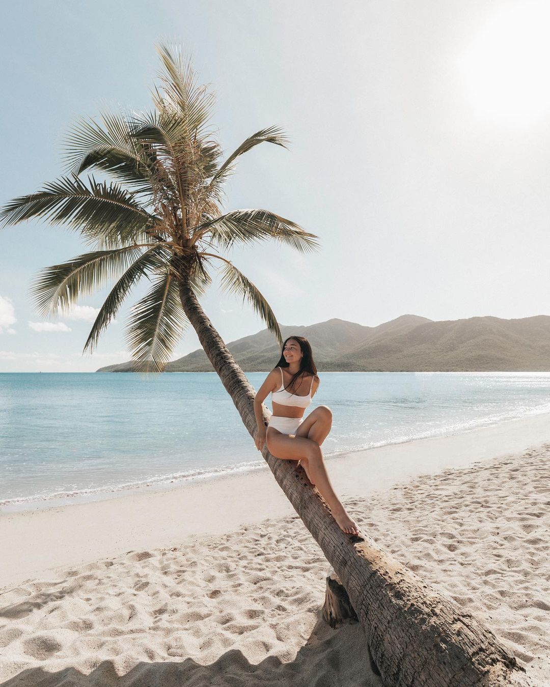 Woman in a white bikini sits on a palm tree at the beach with mountains and ocean in the background on a sunny day.