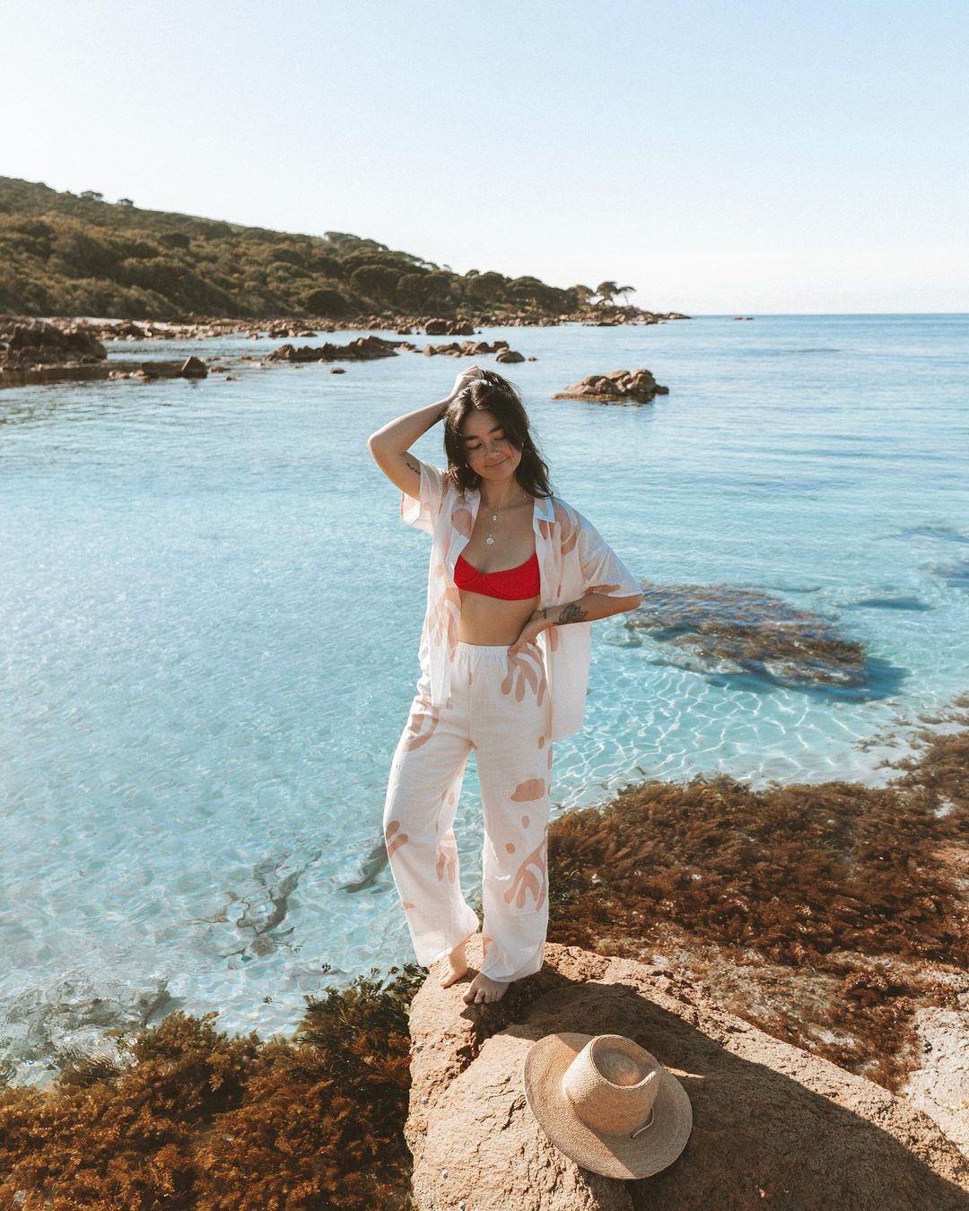 Woman in a patterned shirt and pants stands on a rocky outcrop overlooking clear turquoise water with a hat at her feet.