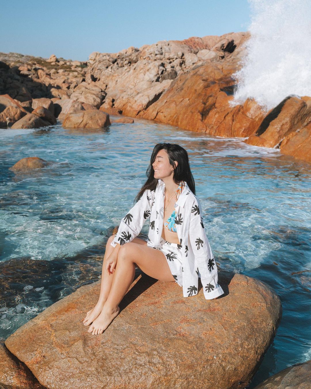 Woman in palm-printed shirt and shorts sits on coastal rock formation near ocean waves, with blue sky and sandstone landscape.