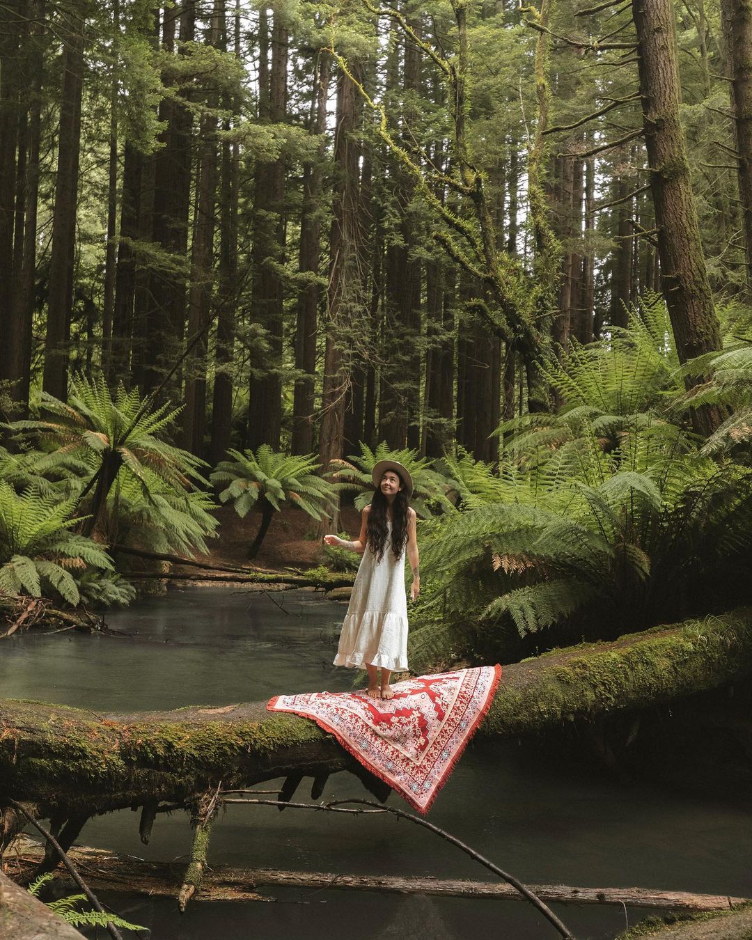 Woman in long dress stands on mossy log with patterned blanket in lush forest by river surrounded by tall trees and green ferns.