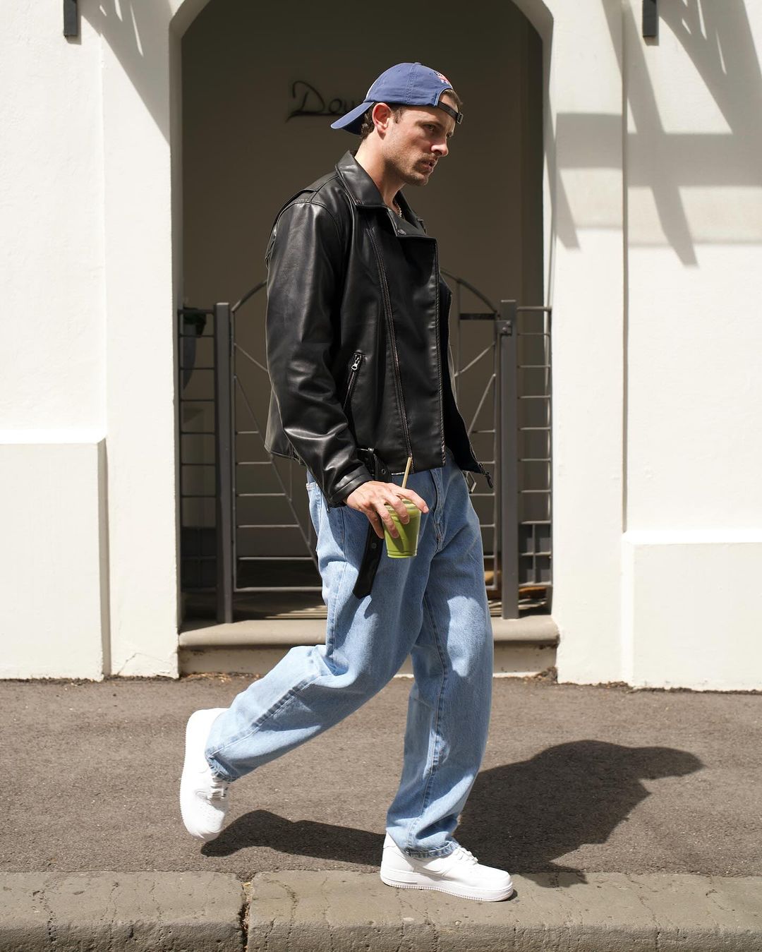 A male adult walks on a sunny street wearing a leather jacket, jeans, sneakers, and a baseball cap, holding a green drink in hand.