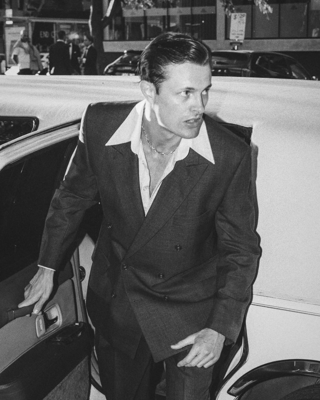 Black and white image of a man in a suit exiting a limo, captured in an urban setting with pedestrians in the background.
