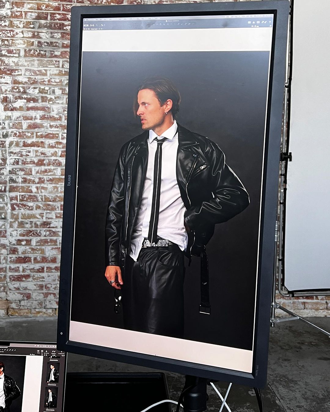 Man in a black leather jacket and tie is displayed on a monitor in a photography studio with a brick wall backdrop. The scene is a photo shoot.