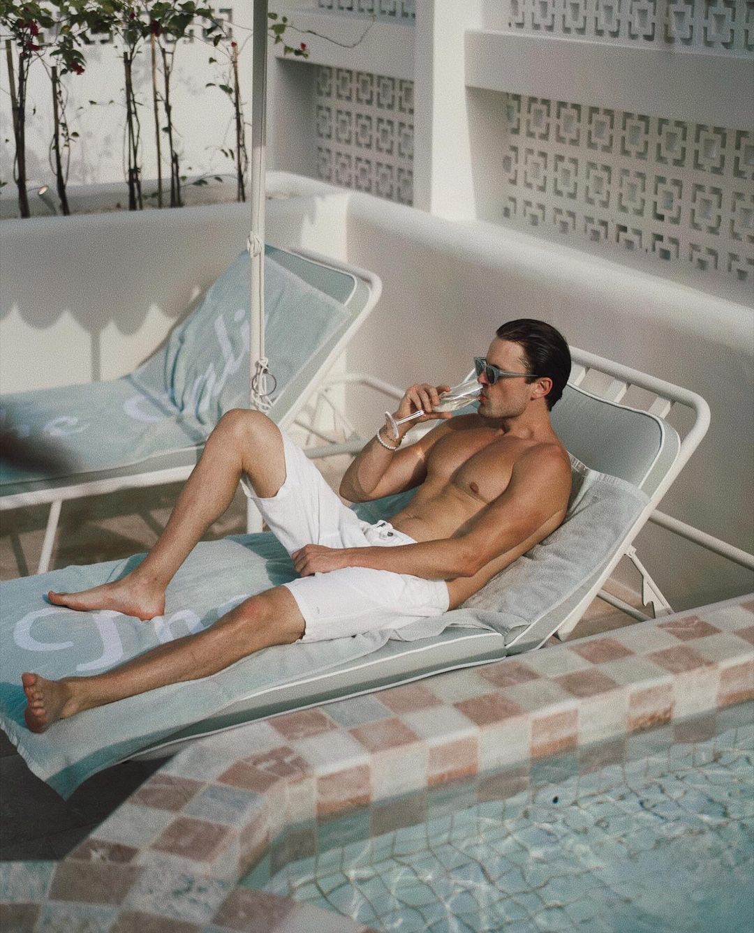 Man lounges topless by the pool, drinking from a champagne flute. He wears white shorts and sunglasses in an outdoor setting.