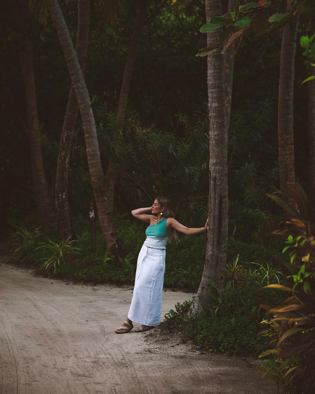 Woman in turquoise top and white skirt poses next to a palm tree on sandy path in tropical forest setting, wearing gold jewelry.