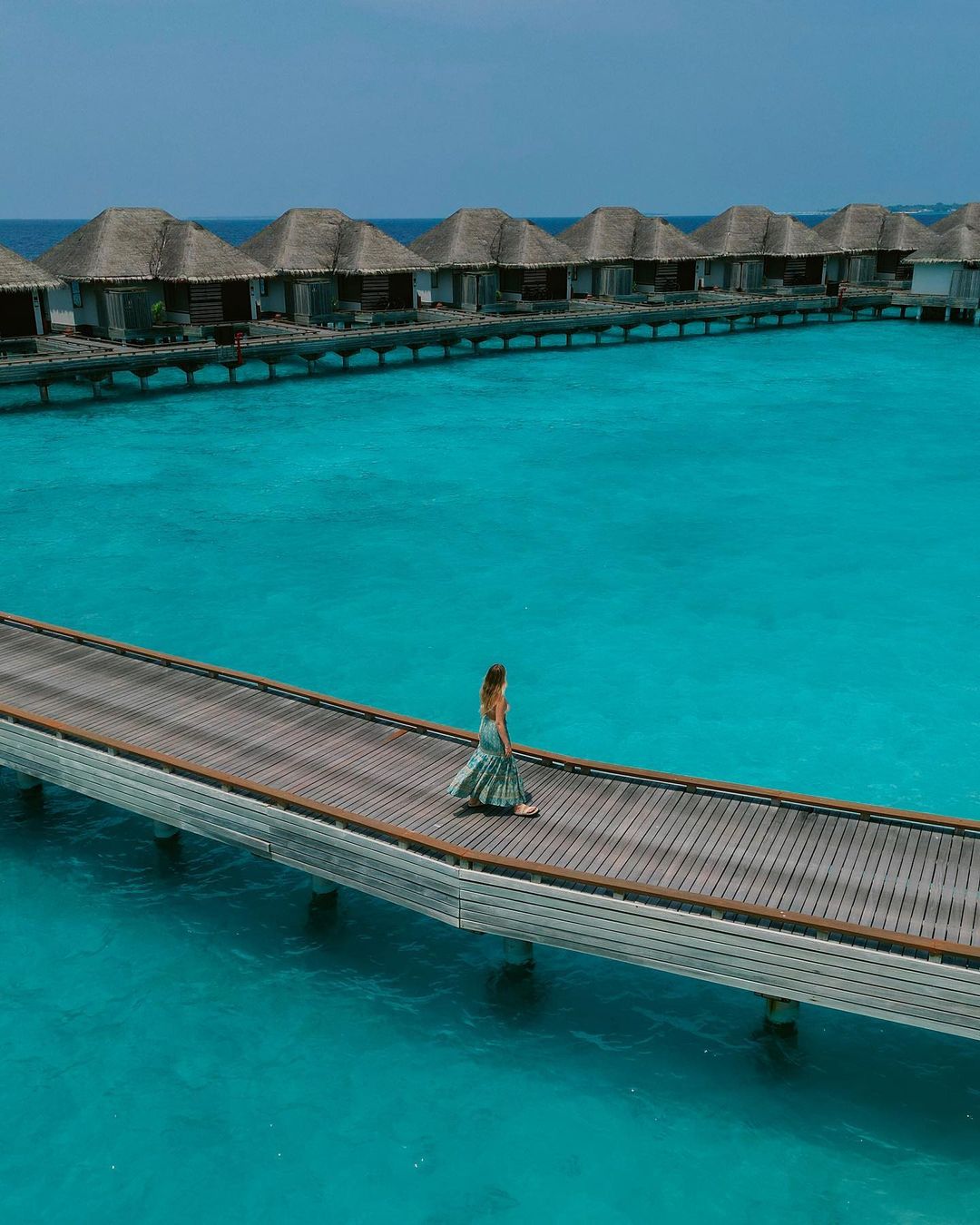 Woman in a long patterned dress walks along a wooden pier towards thatched-roof bungalows over turquoise water on a sunny day.