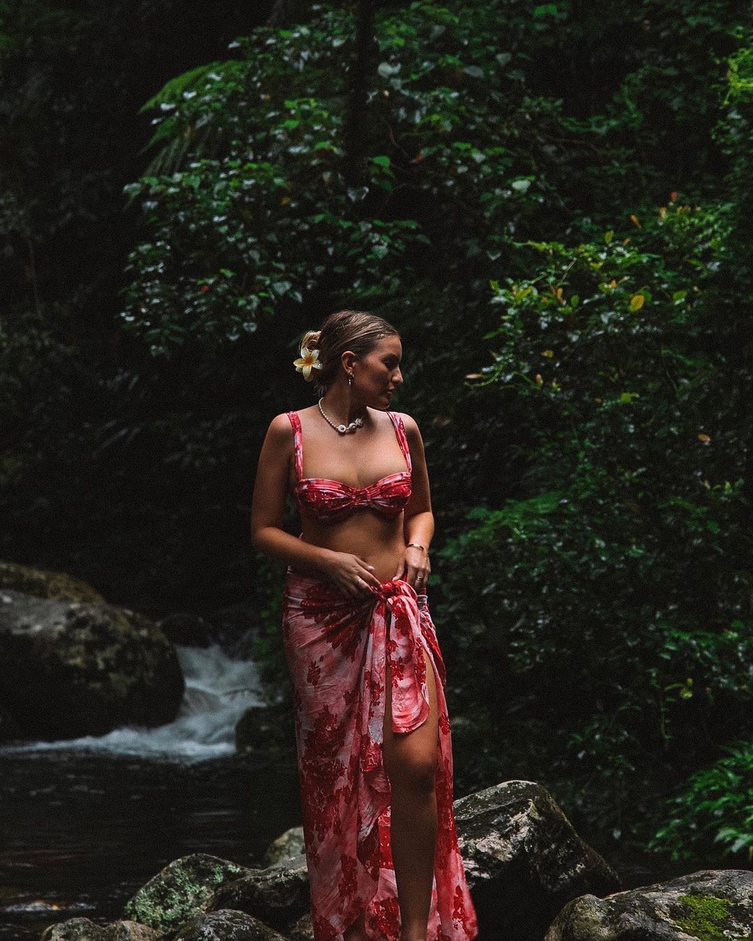 Woman in floral bikini and wrap stands on rocks near waterfall in lush rainforest setting; tropical vacation or outdoor fashion shoot.