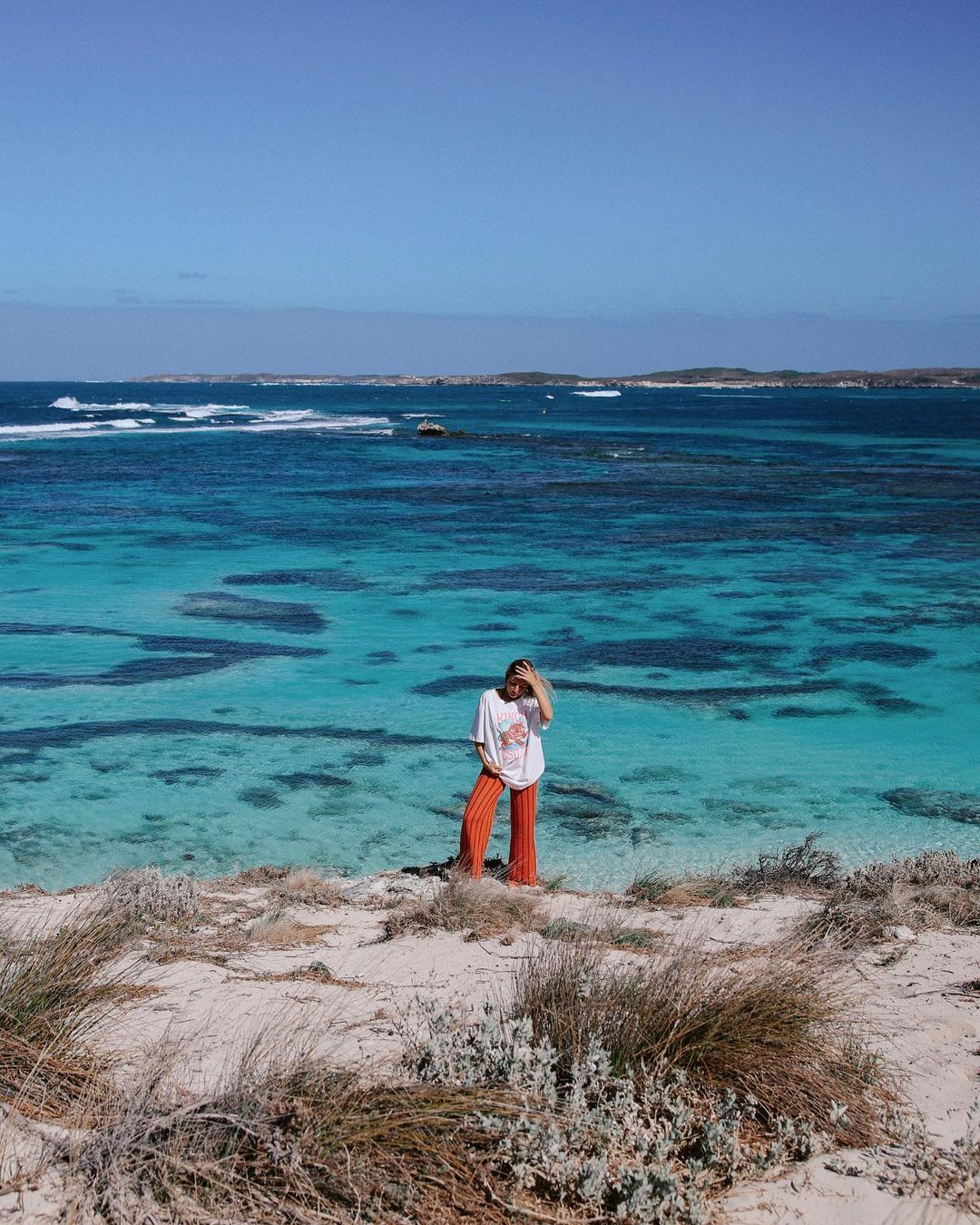 A woman in orange pants and a white shirt stands on a sandy dune overlooking turquoise water under a clear blue sky in a scenic outdoor shot.
