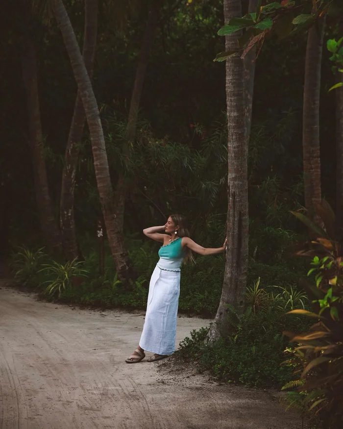 Woman in green one-shoulder top and white skirt leaning against a palm tree on a sandy pathway by Aimee Bell.