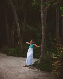 Woman in green one-shoulder top and white skirt leaning against a palm tree on a sandy pathway by Aimee Bell.