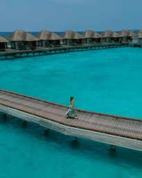 Aerial view of a woman walking on a wooden jetty over turquoise ocean water toward a row of thatched-roof overwater bungalows.