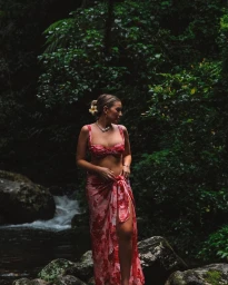 Aimee Bell wearing a red floral bikini and matching sarong standing outdoors near a rocky stream in a lush green forest.