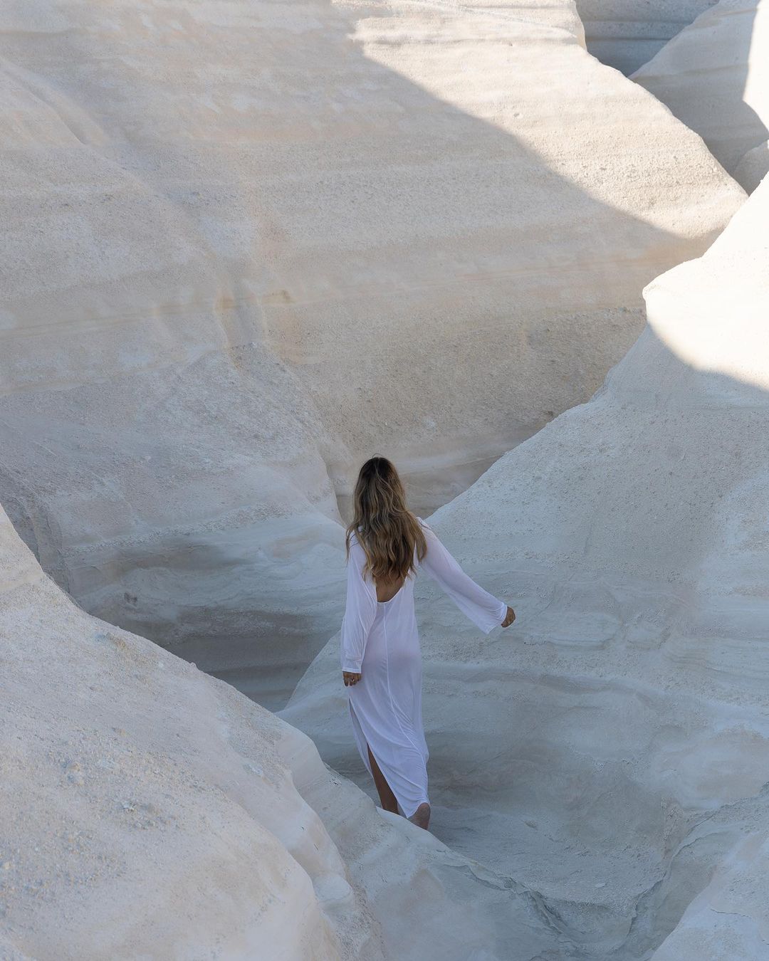 A woman with long brown hair wearing a long white dress walks through a white rock canyon. The light is bright, casting shadows on the stone.