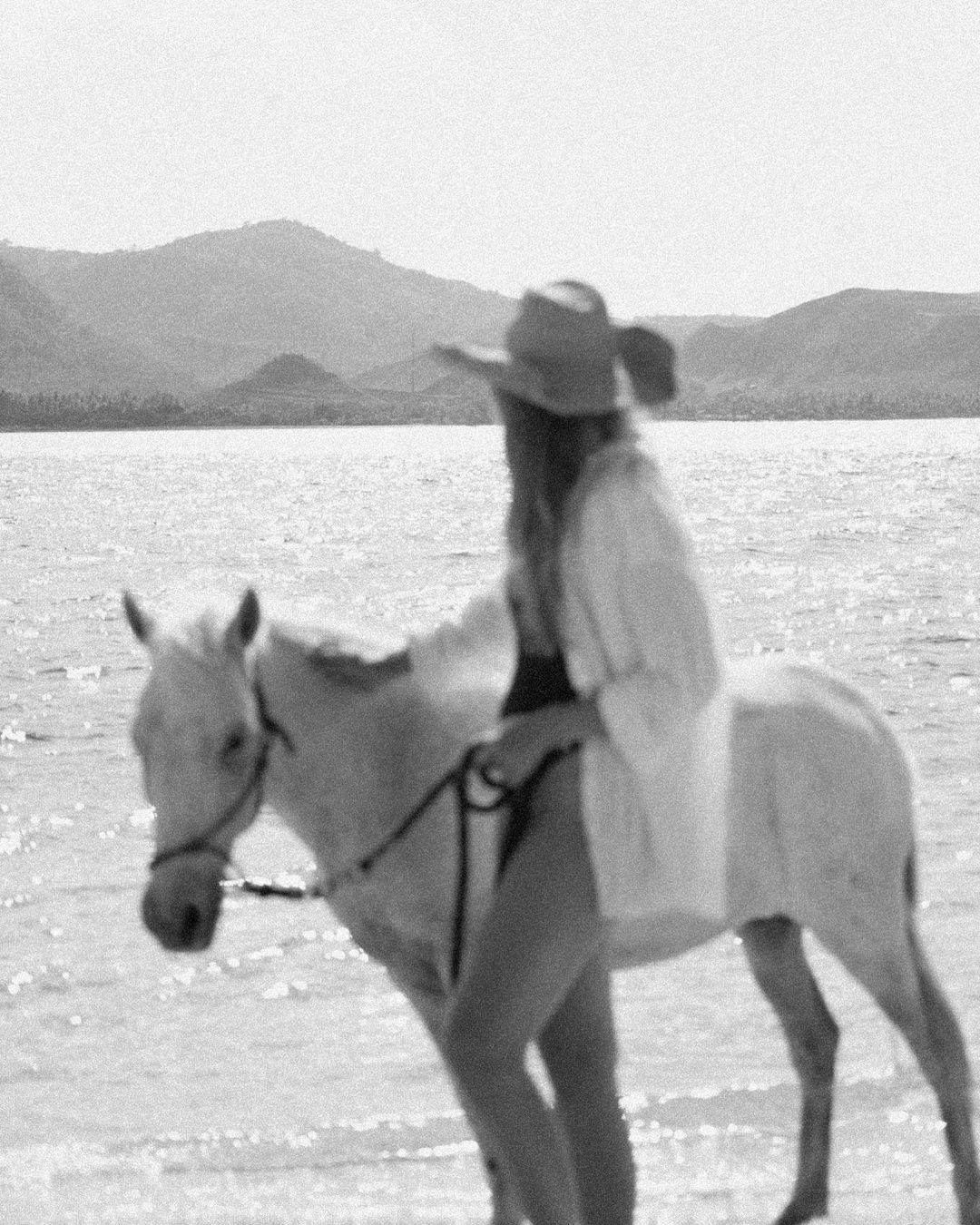 Woman riding a white horse on a beach with ocean and mountains in the background, black and white outdoor shot.