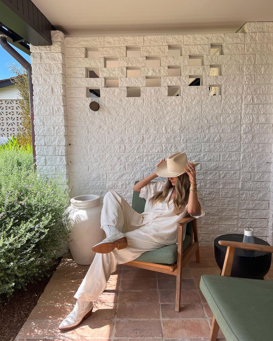 Adult female in neutral-tone clothing and hat reclines in a wood chair on a patio with brick flooring and a white-brick background wall.