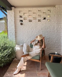 Woman wearing cream cowboy hat and white outfit seated in patio chair against white brick wall