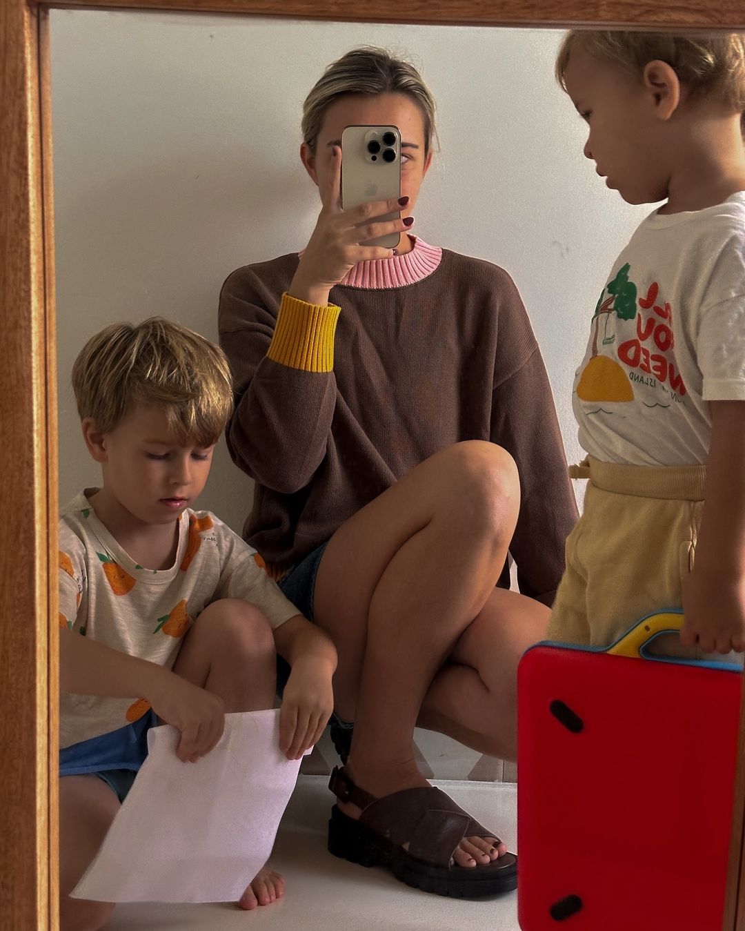 A woman in a brown sweater takes a selfie in the mirror with two young children standing and sitting beside her in an indoor setting.