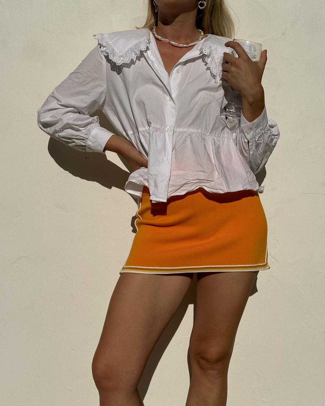 Woman poses in a white, frilled blouse and short orange skirt in front of a light-colored wall, showcasing a fashion forward look.