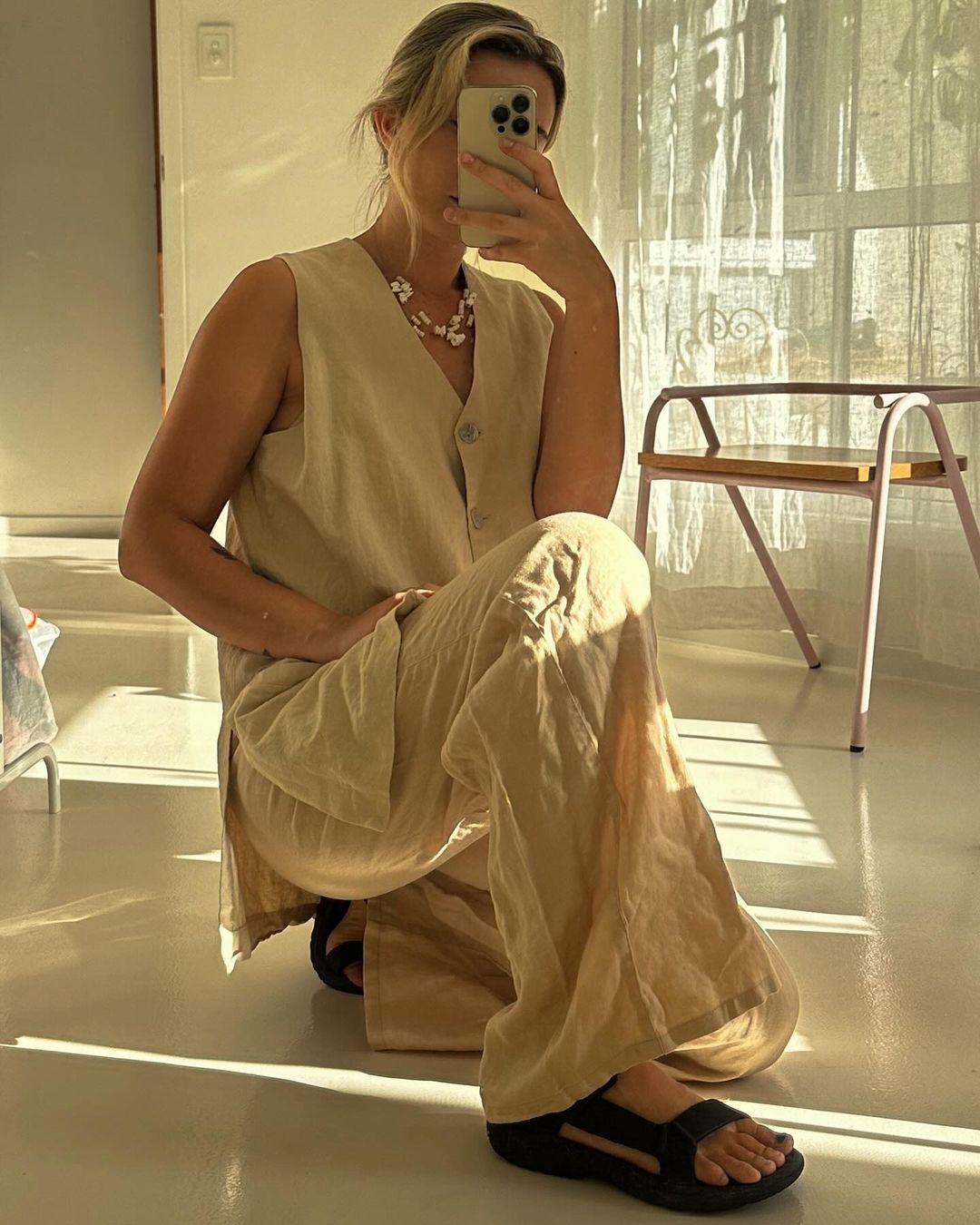 Blond woman takes a mirror selfie, kneeling indoors wearing a vest and pants with sandals. Natural light streams through the room.