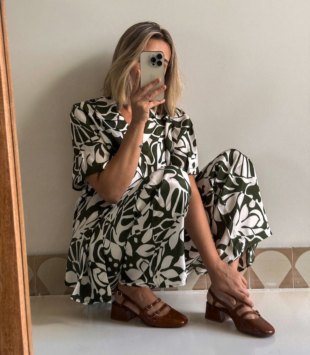 A woman is crouching indoors wearing a patterned green and white jumpsuit, taking a photo of herself with a smartphone, with a neutral background.