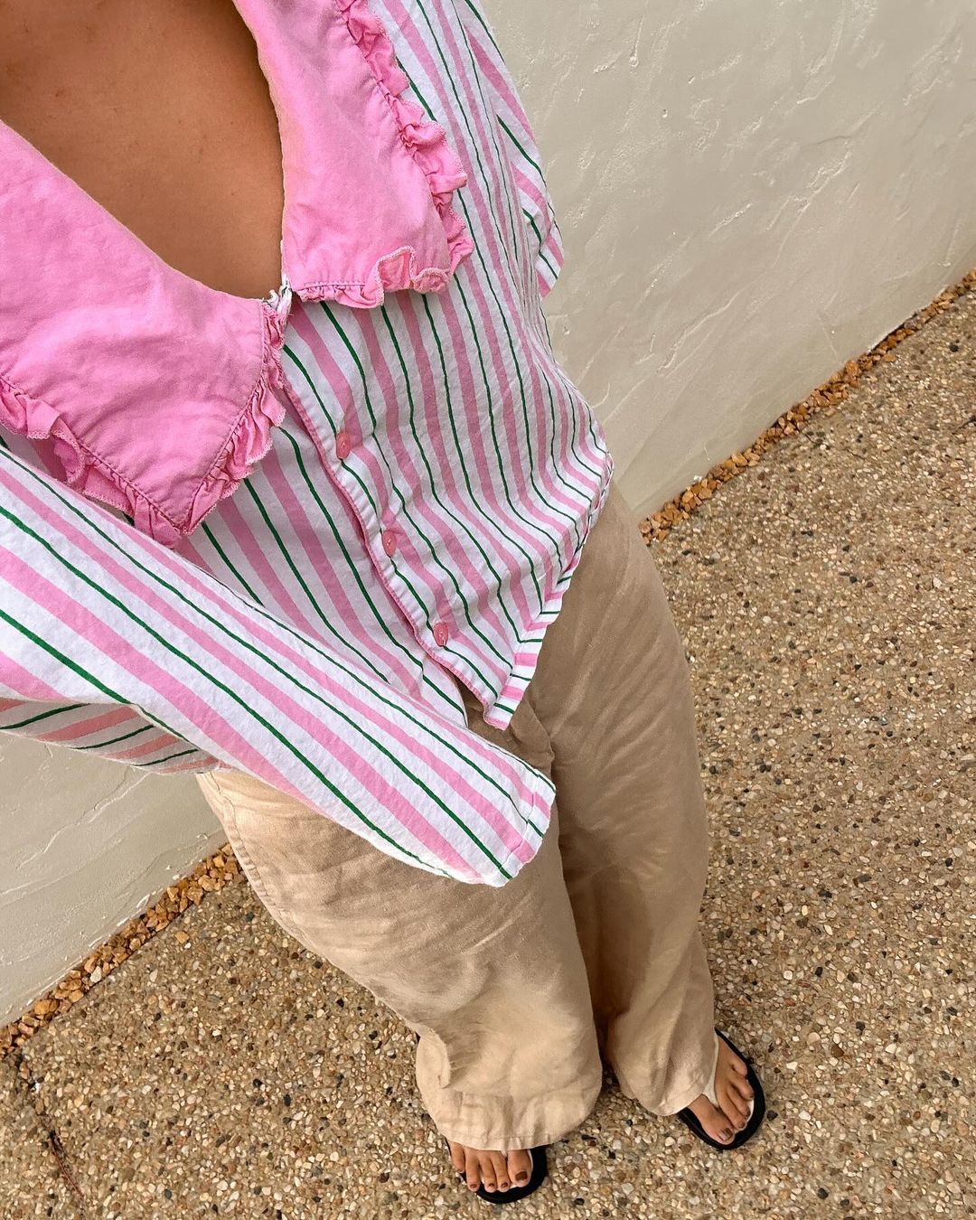 A person wears a pink and white striped collared shirt with beige trousers and black sandals standing on a gravel and stone surface.