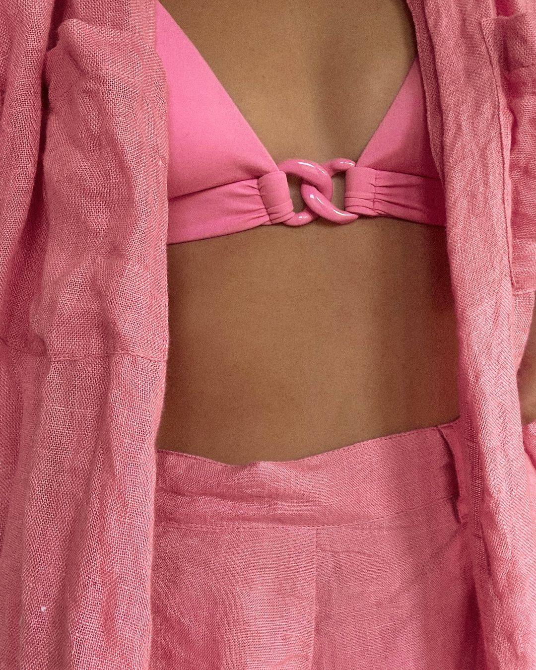 Close-up of a woman's torso wearing a pink bikini top and an open linen shirt with matching pink linen pants.