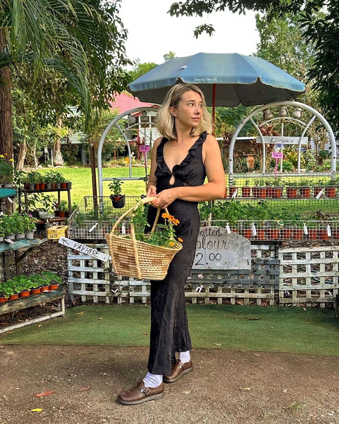 A woman wearing a black dress and carrying a basket with flowers in a garden setting, surrounded by greenery and potted plants.