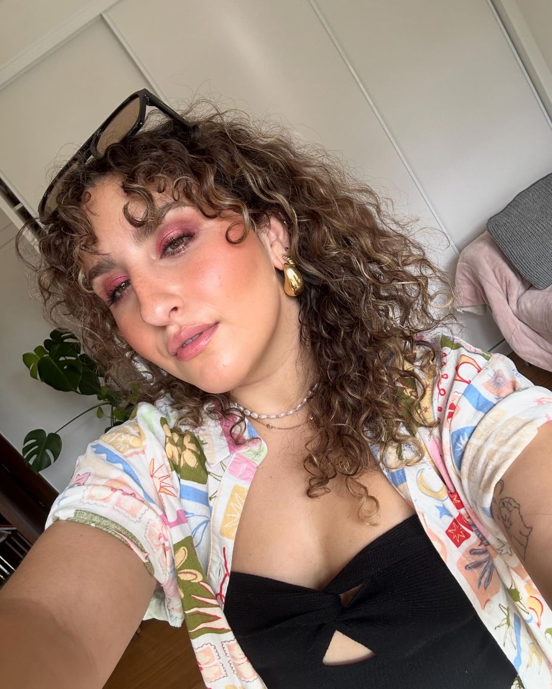 Woman with curly hair, wearing a patterned shirt and black top, poses indoors, showcasing her makeup and jewelry in a close-up selfie.
