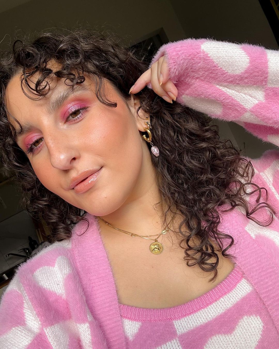 Close-up of an adult female with curly hair, wearing a pink and white patterned sweater, jewelry, and pink eyeshadow indoors.