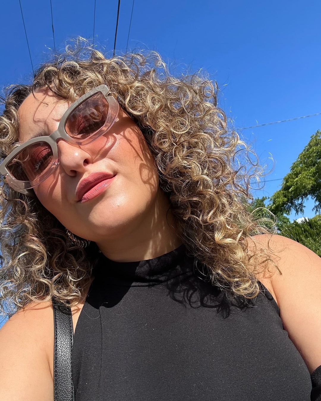 A woman with curly hair and sunglasses looks upward in an outdoor shot. The focus is on her face, hair and fashion.