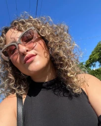 Close-up portrait of a woman with curly blonde hair and sunglasses wearing a black top against a clear blue sky by Clary Riven.