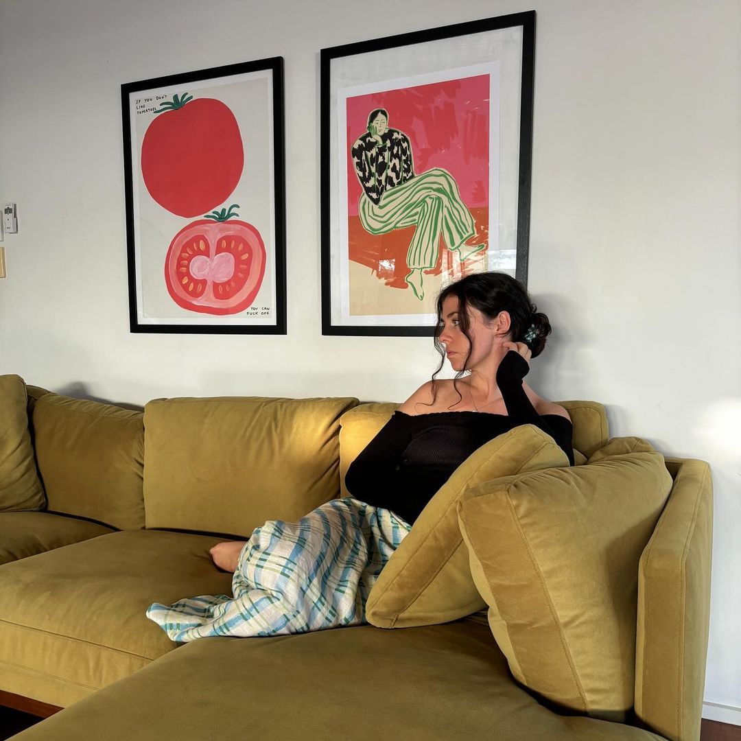 Woman relaxes on a mustard-colored sofa in a warmly lit living room, with colorful art on the wall, and a cozy, domestic atmosphere.