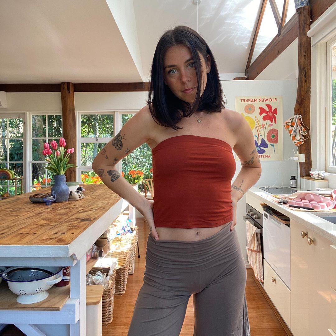 Woman in rust-colored top and grey pants stands in a light-filled kitchen with wood beams, tulips, and a colorful flower market poster.