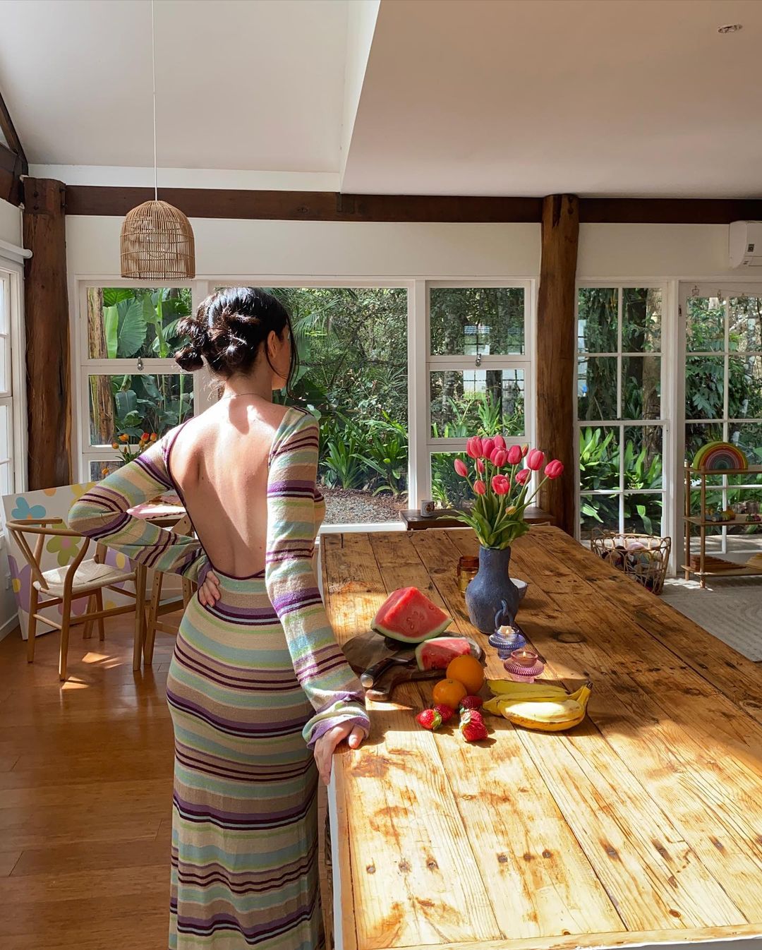Woman in striped dress in sunlit kitchen with wooden table, tulips, and fruit, enjoying a bright day at home.