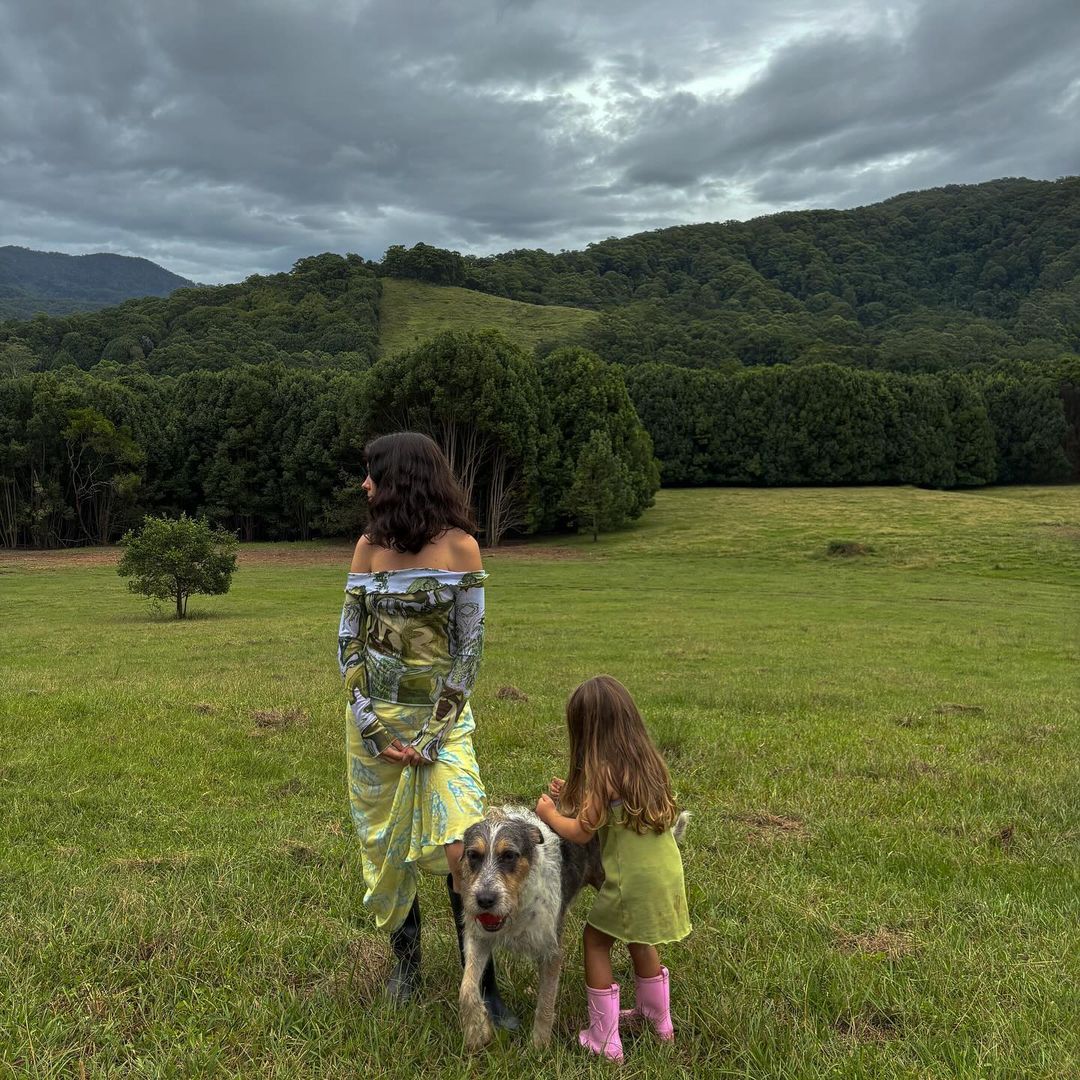 Woman and child with a dog in a grassy field, with rolling hills and a cloudy sky. The woman wears a landscape-printed dress.