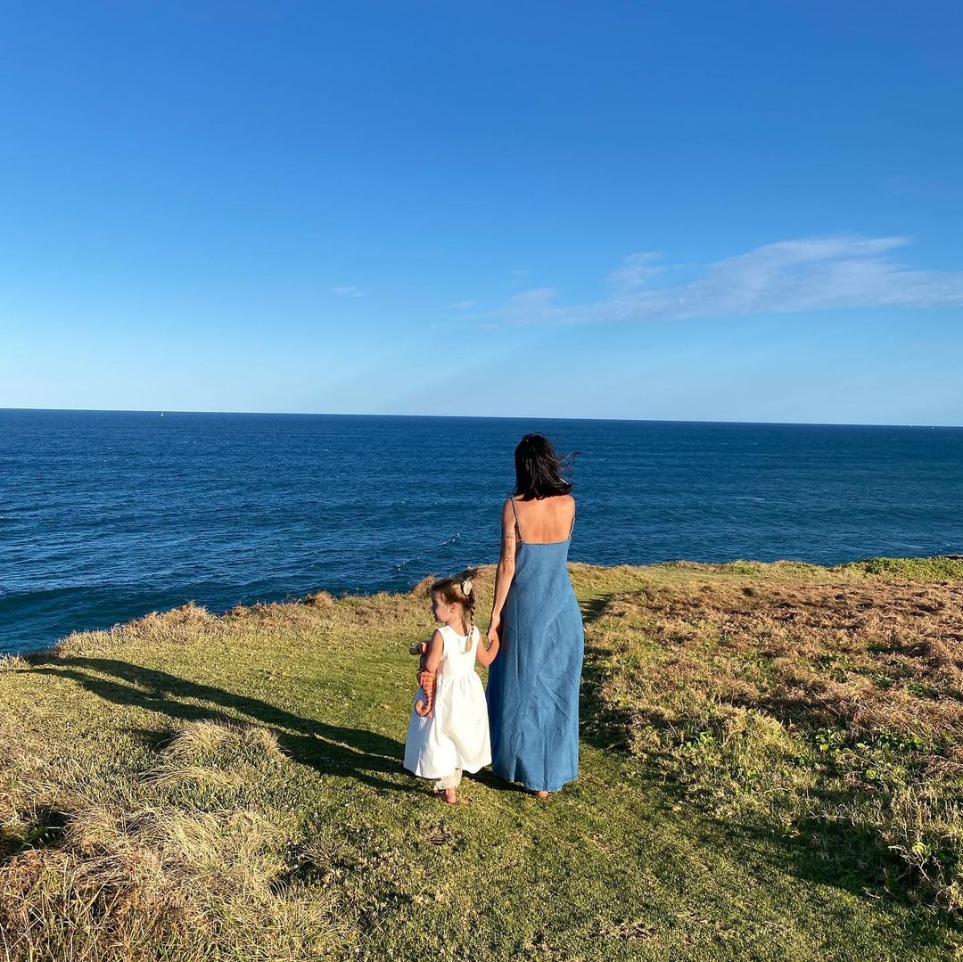 A young girl and an adult female holding hands, stand on a grassy hill overlooking the ocean under a clear blue sky on a sunny day.