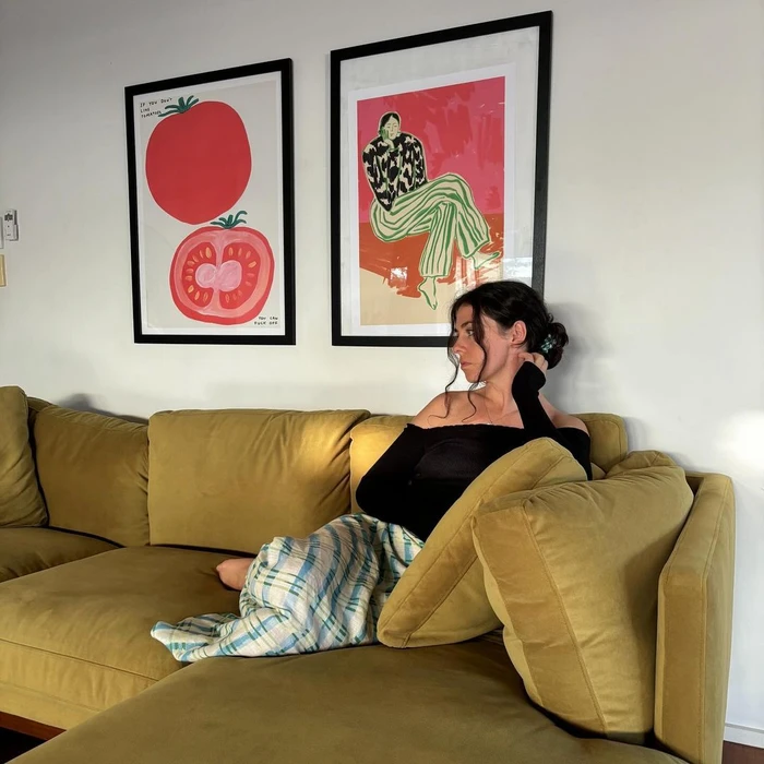 Woman in black off-shoulder top sitting on a mustard yellow sectional sofa with framed art prints on the wall behind.