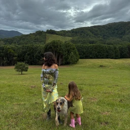 Woman and young child standing with a shaggy dog in a grassy meadow under a cloudy sky.
