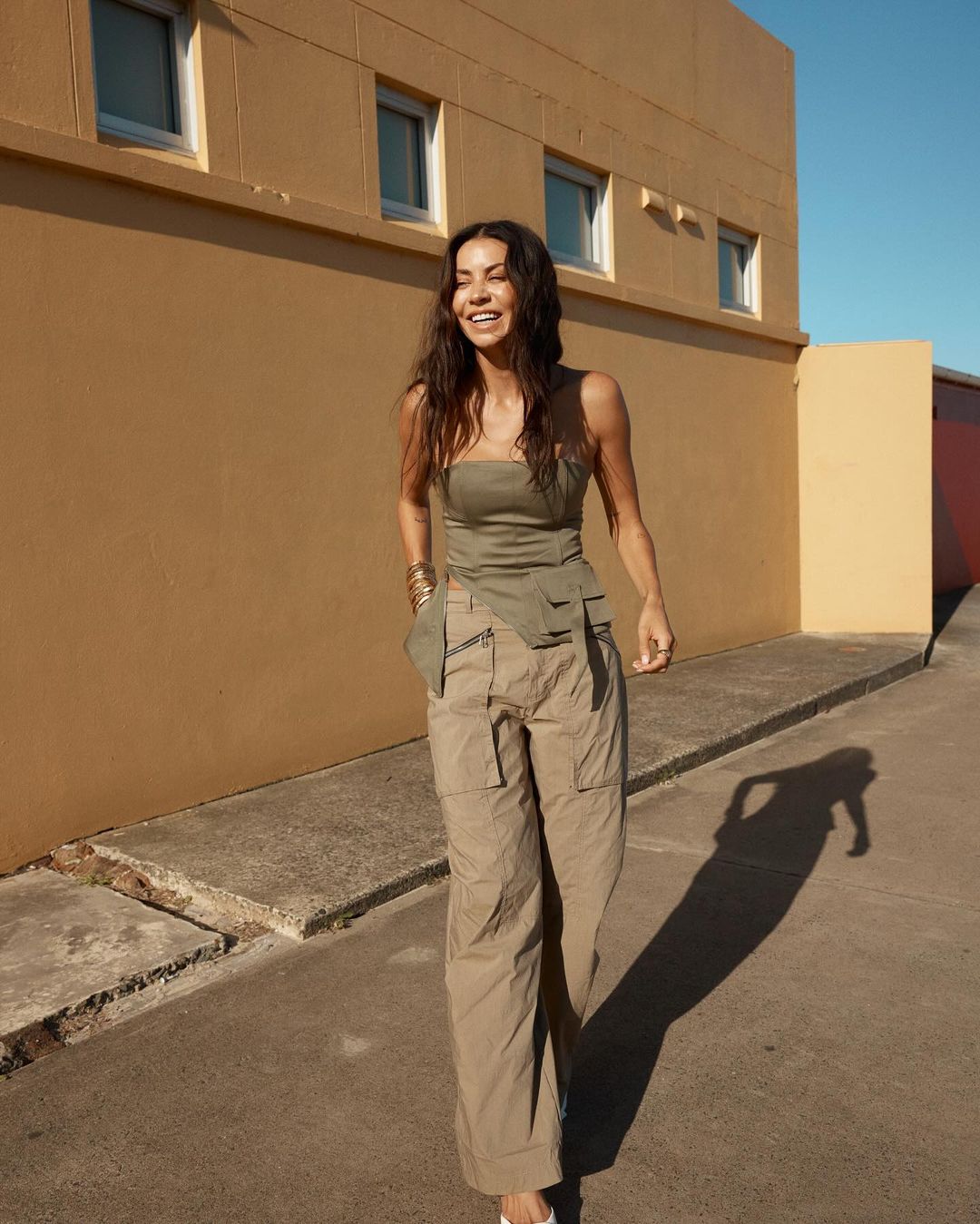 A woman smiles and walks down a paved path next to a tan-colored building wearing a strapless top and beige cargo pants outdoors on a sunny day.