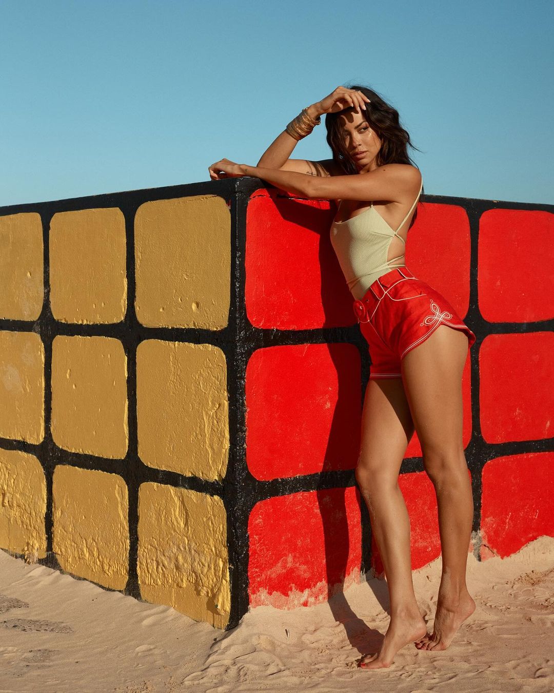 Woman poses barefoot in red shorts and a top near a large, colorful cube on a sandy surface beneath a clear, blue sky.
