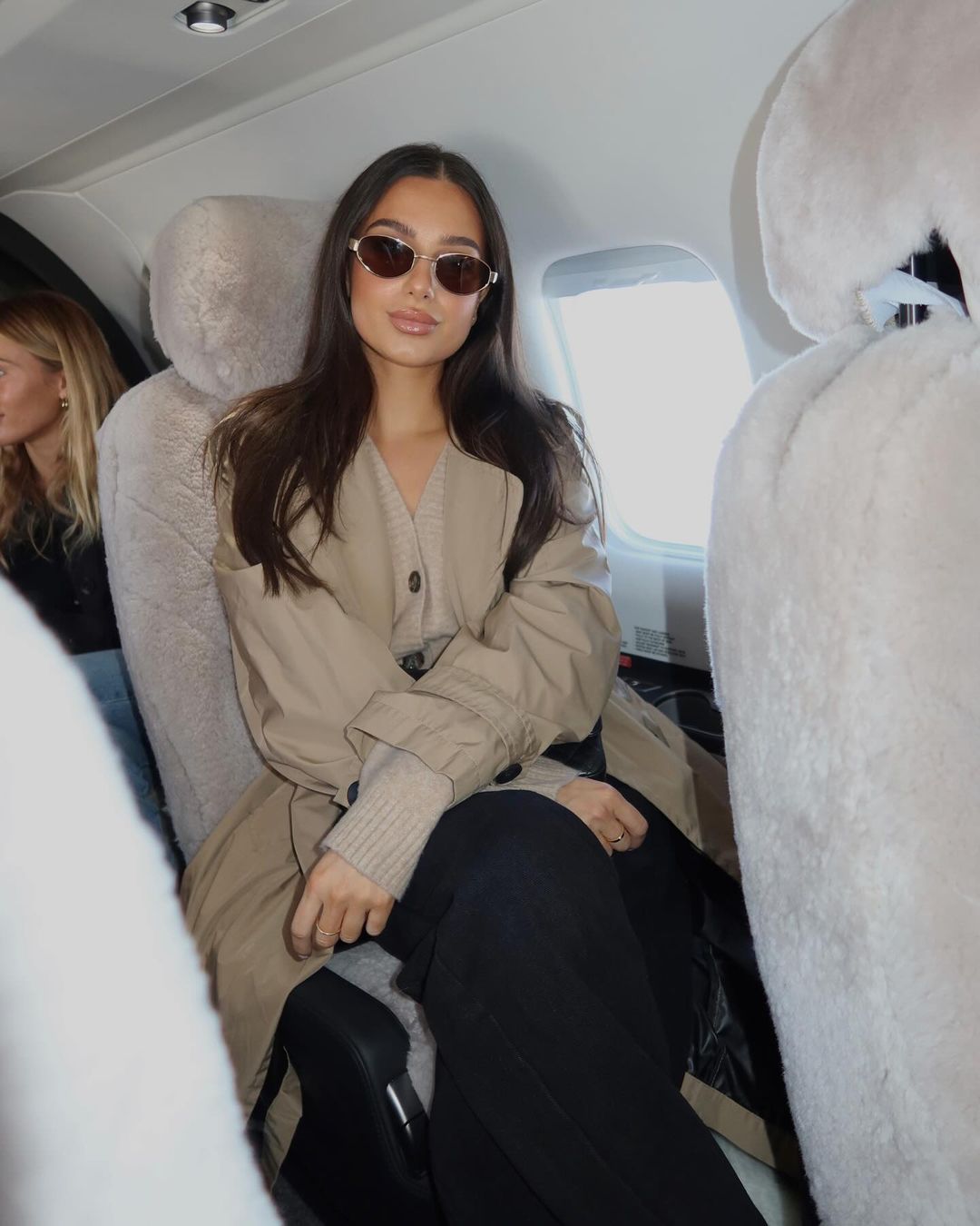 Attractive woman with sunglasses sits on an airplane wearing a coat, creating a stylish travel moment in a private aircraft.