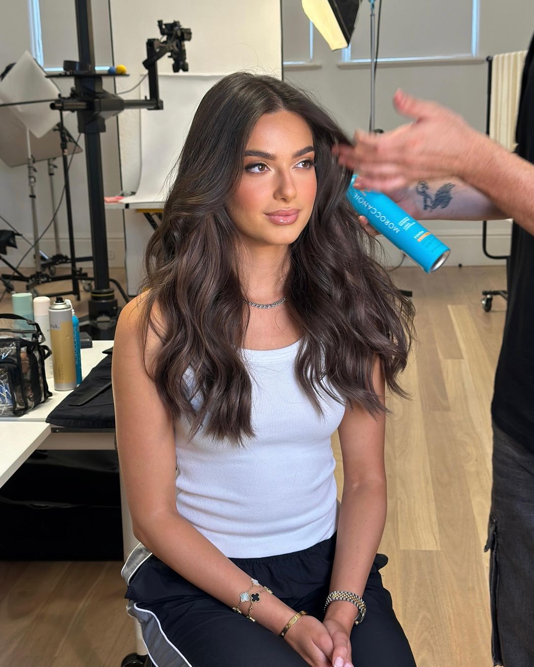 Brunette woman with styled wavy hair poses while getting hairspray applied in a bright photo studio with lighting equipment visible.
