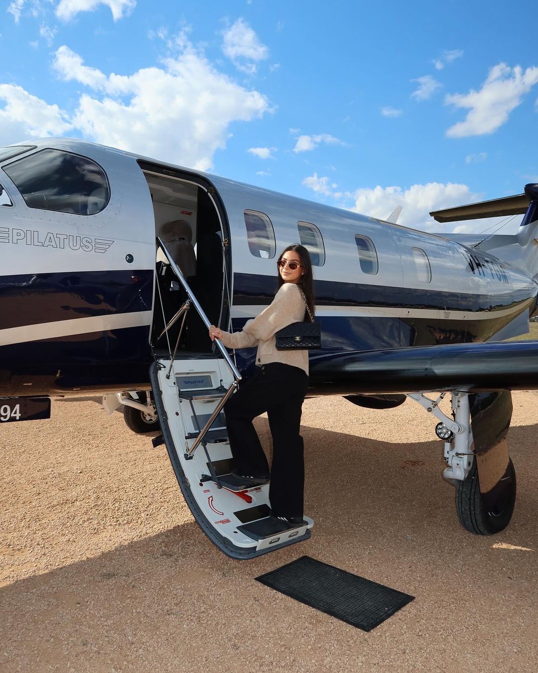 Woman boards private jet wearing sunglasses and black slacks, under a sunny blue sky with wispy clouds, travel, lifestyle, and fashion.