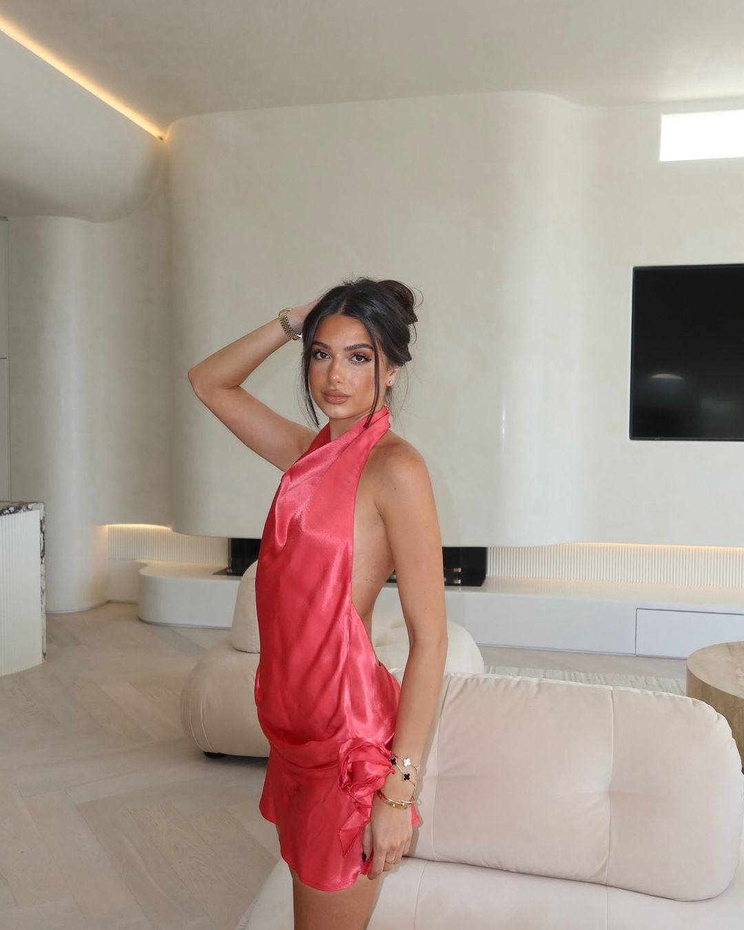 Woman in a coral-colored, halter-neck mini dress poses indoors. The dress has a draped design. She wears jewelry and has an updo hairstyle.