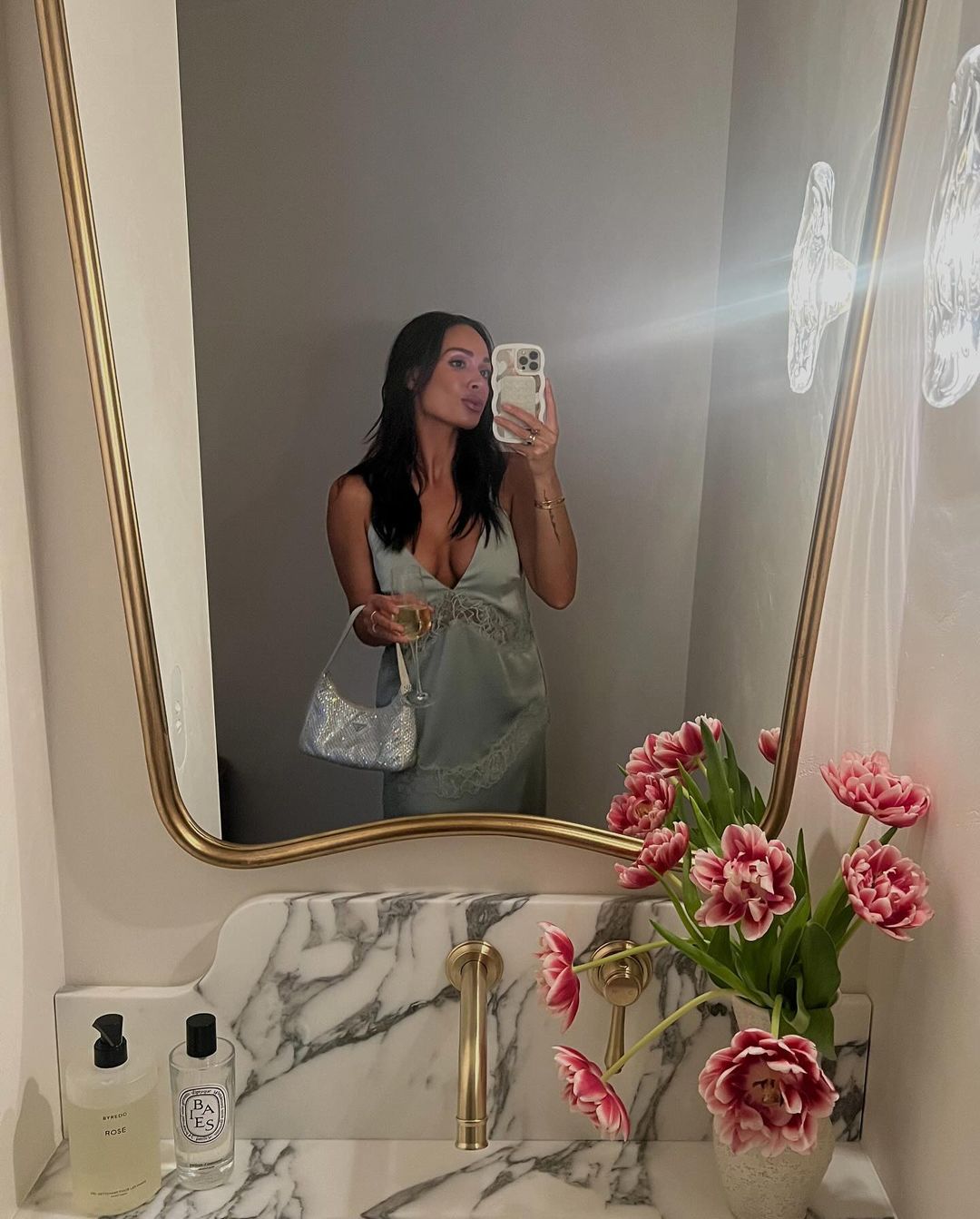 Brunette woman in a green satin dress takes a mirror selfie in a bathroom with pink tulips, a marble counter, and gold fixtures.