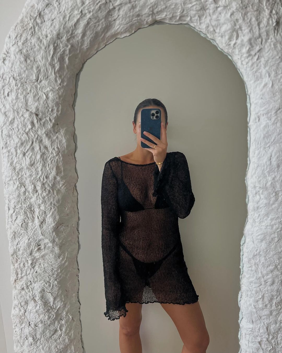 Young adult woman takes a mirror selfie in a black swim suit and a black sheer cover-up dress in an indoor environment with neutral tones.