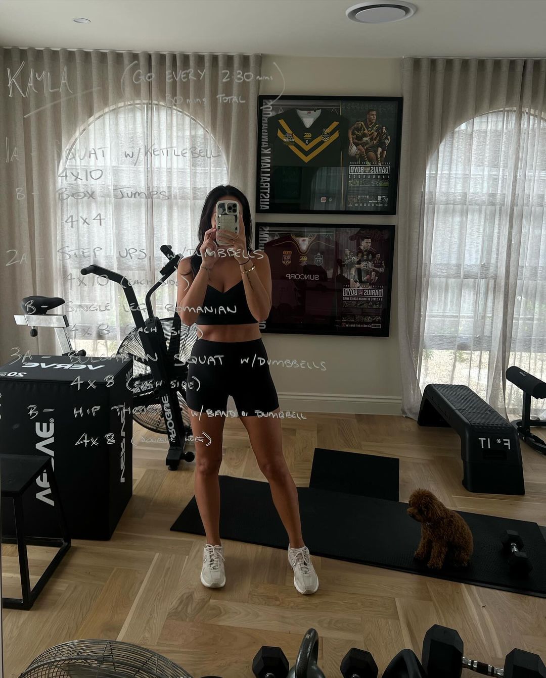 A woman in workout clothes stands in a home gym with exercise equipment, a mirror with workout notes, and a small dog on an exercise mat.